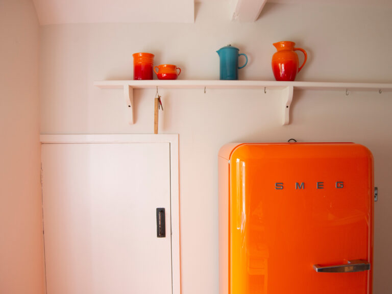 Bright orange SMEG fridge against a white wall with a small shelf above with colourful jugs.