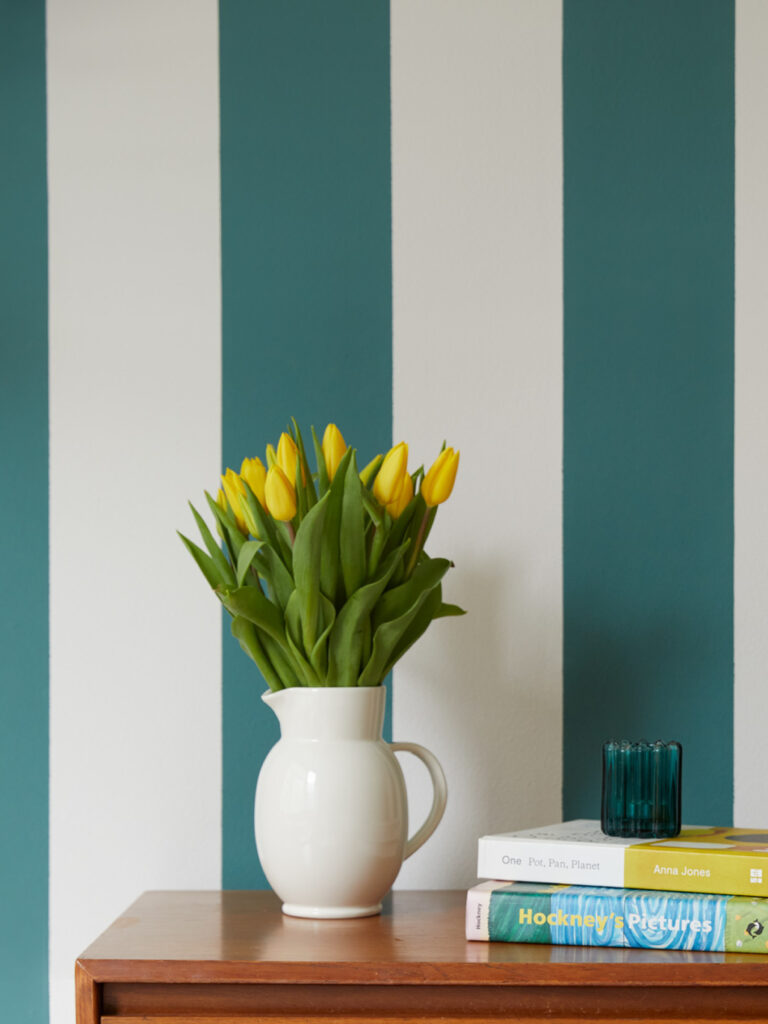 A striped wall with a table and a jug full of fresh yellow tulips.
