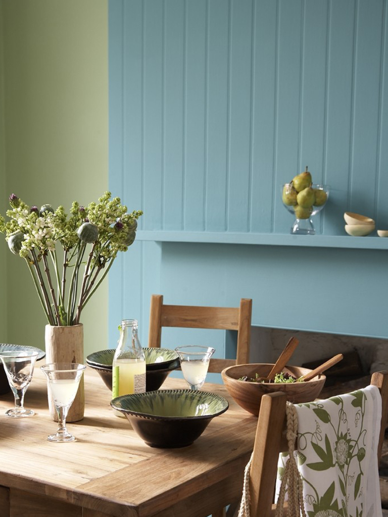 Wooden kitchen table setting with a green wall behind that has blue panelling around a fire place. Fresh summer time.
