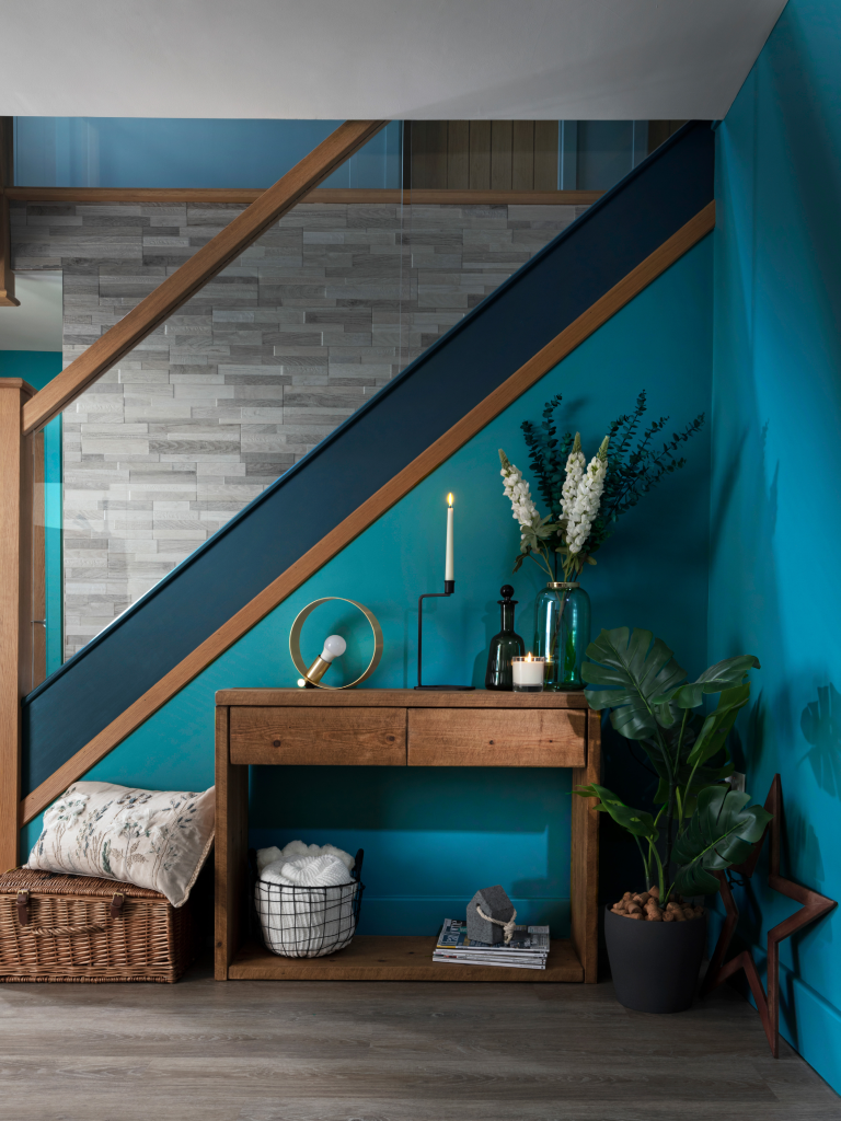 Side profile of a stair case with vibrant blue walls, wooden furniture and a pot plant and vase of flowers.