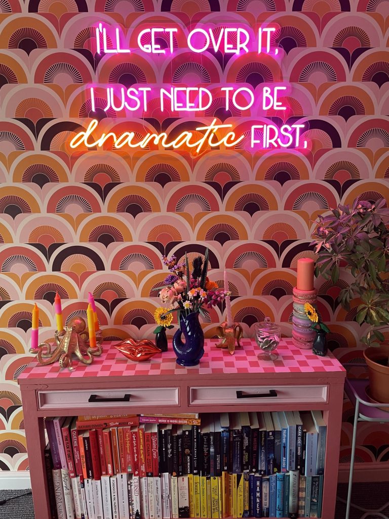 Maximalist style décor of a brightly patterned wall behind a checker painted table stacked with books, candle holders and a vase of flowers.