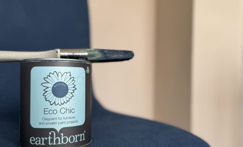 A tin of Earthborn Eco Chic and a used paintbrush on a fabric chair that's been painted with Eco Chic in the colour Balloon Ride.